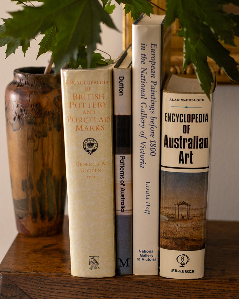 Three books on a wooden surface with a vase and plant in the background
