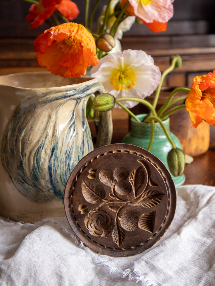 Wooden butter stamp with floral design in front of a ceramic pitcher and flowers.