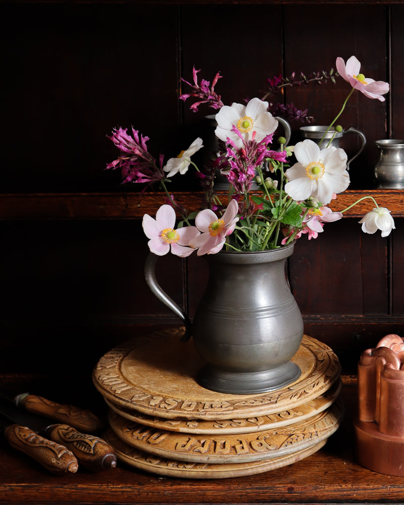 Floral arrangement in a pewter measure on antique breadboards with a dark background