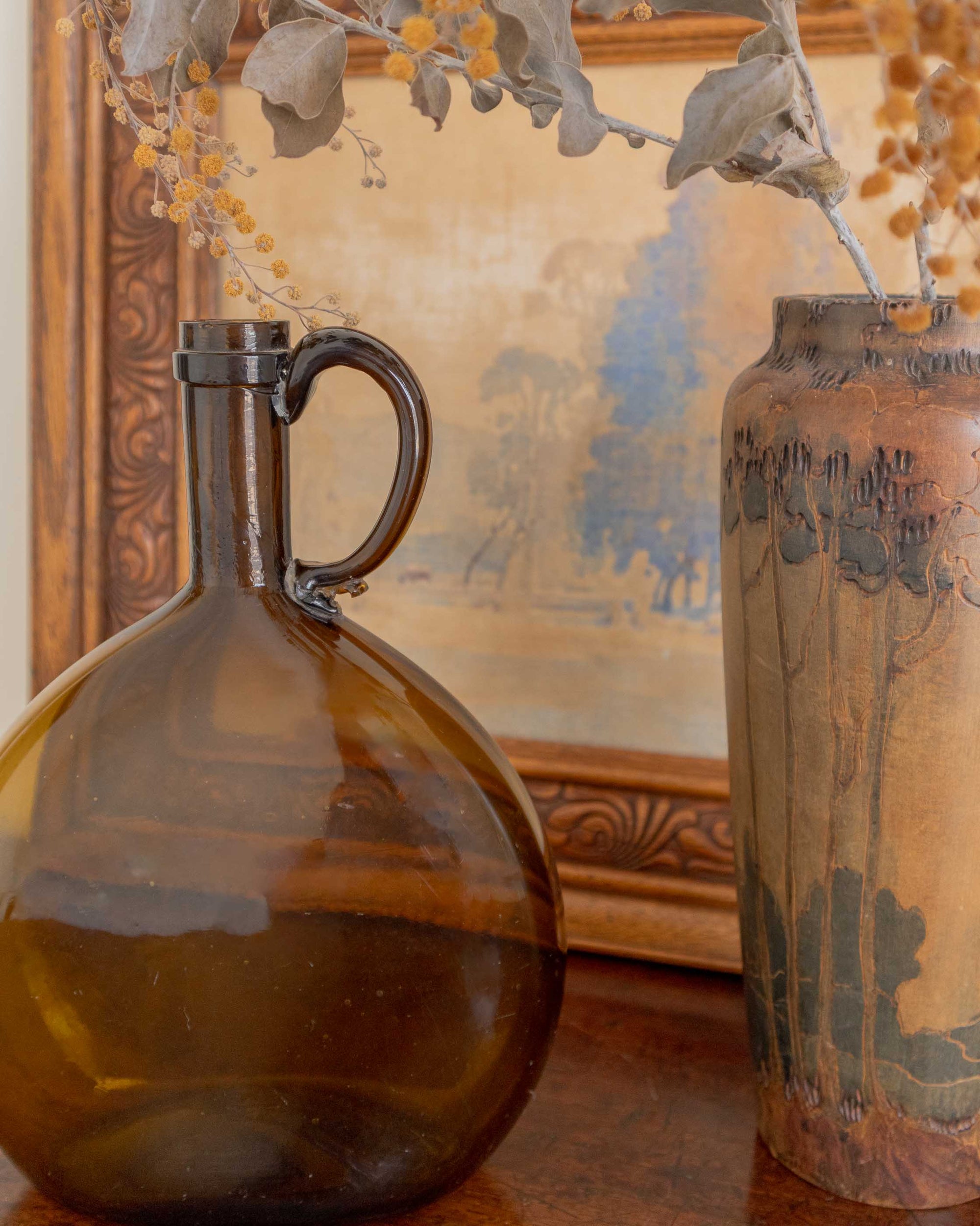 Amber glass flagon on a wooden surface with a decorative painting in the background.