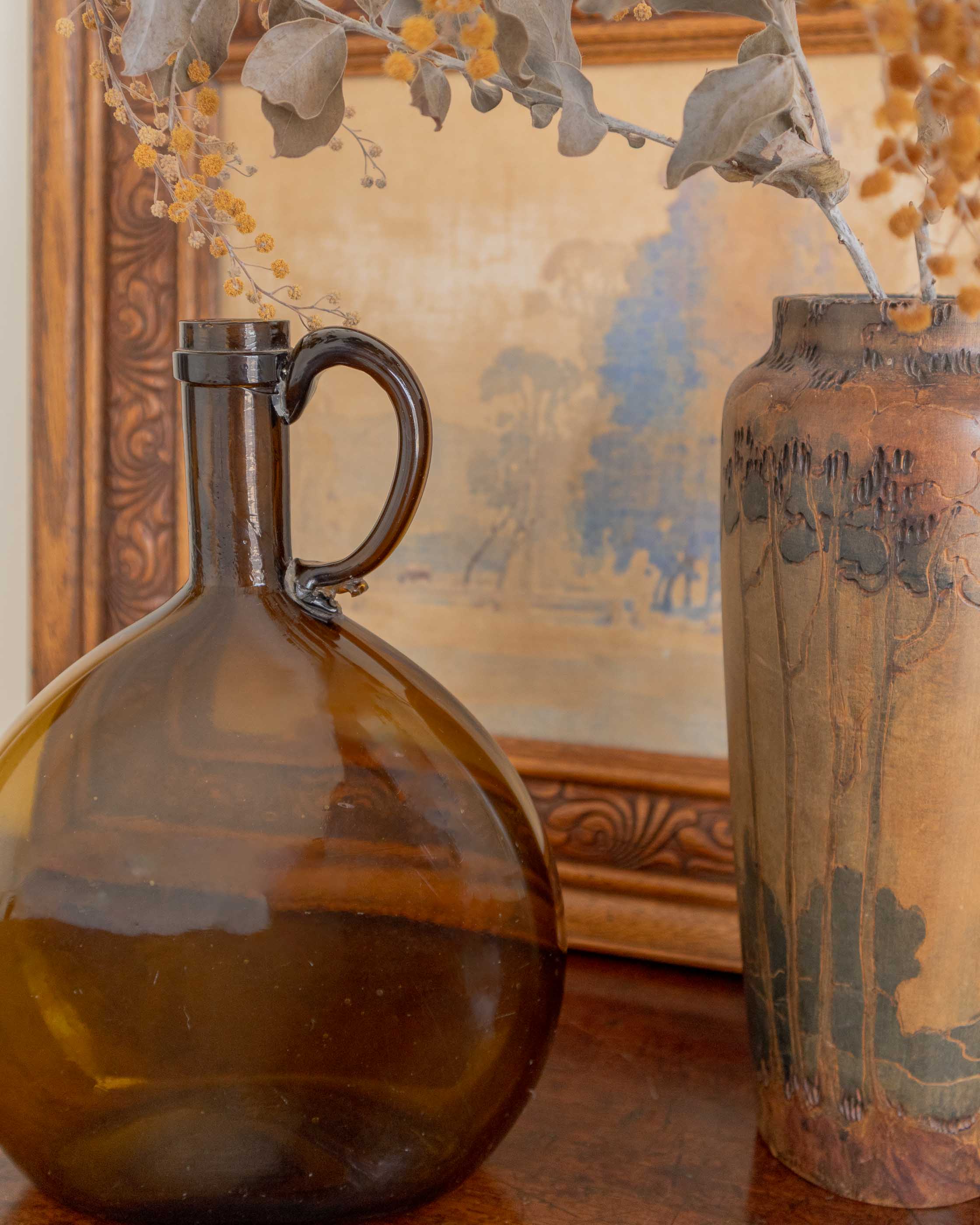 Amber glass flagon on a wooden surface with a decorative painting in the background.