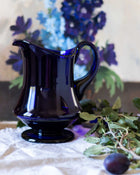 Blue glass pitcher on a white surface with flowers in the background