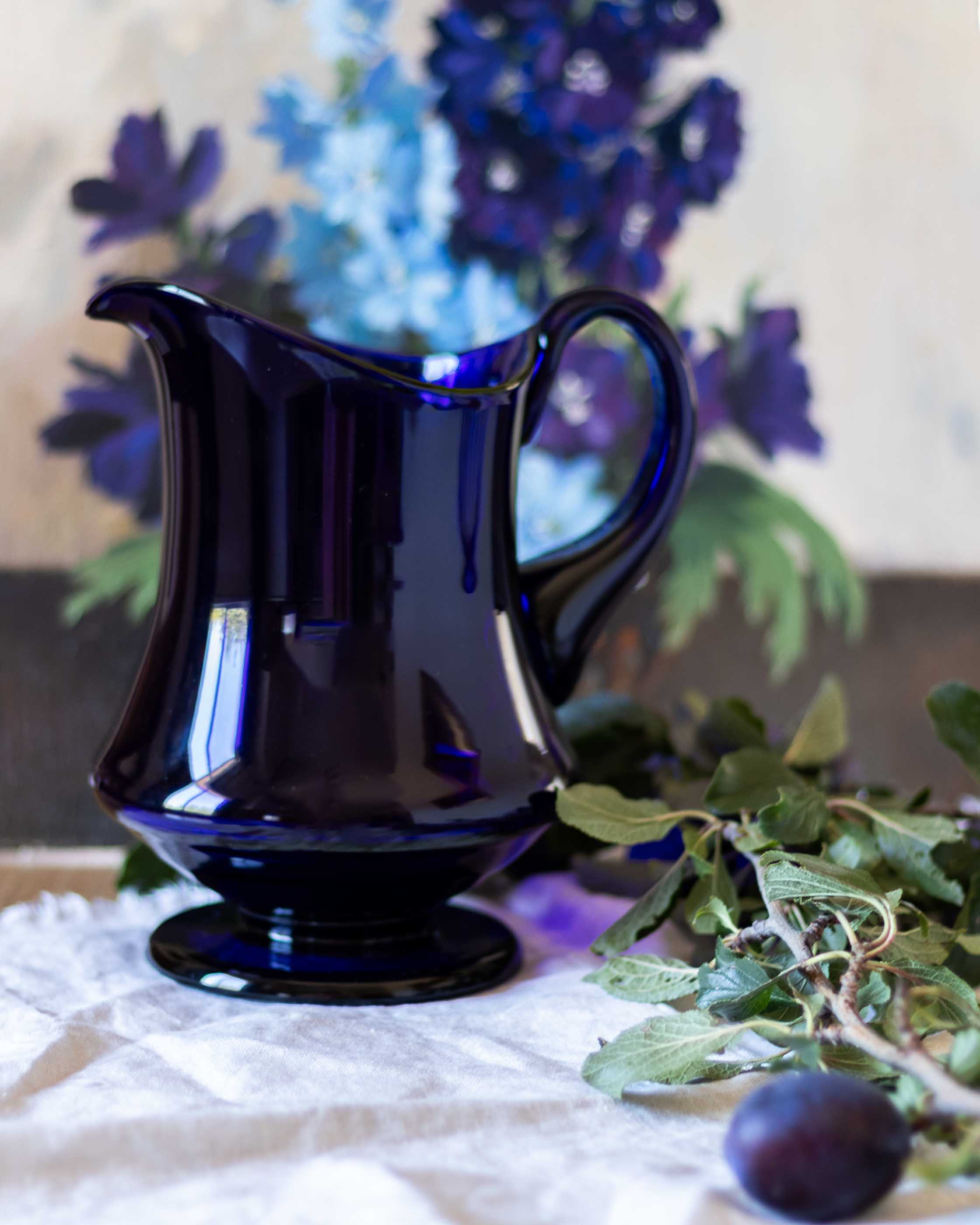 Blue glass pitcher on a white surface with flowers in the background