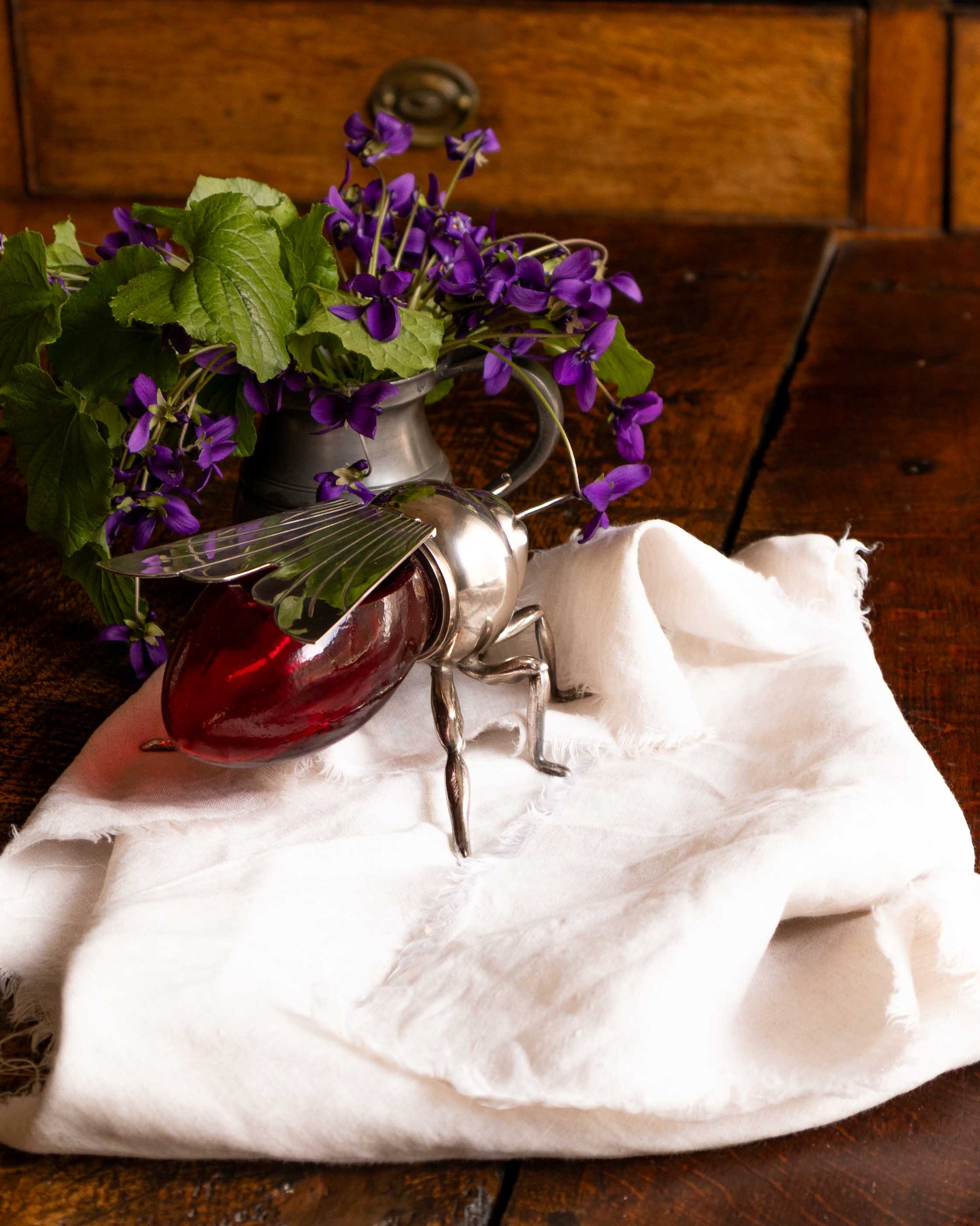 Decorative bee honey pot with a red glass base and metal legs on a wooden surface with flowers in the background