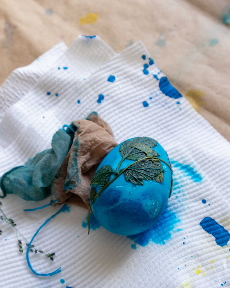 Blue decorative egg with greenery on a white cloth with blue paint splashes