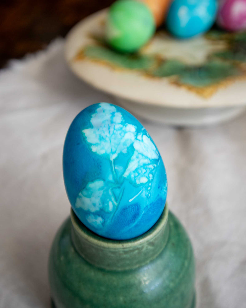 Blue Easter egg with white floral design on a green stand, with more eggs in the background.