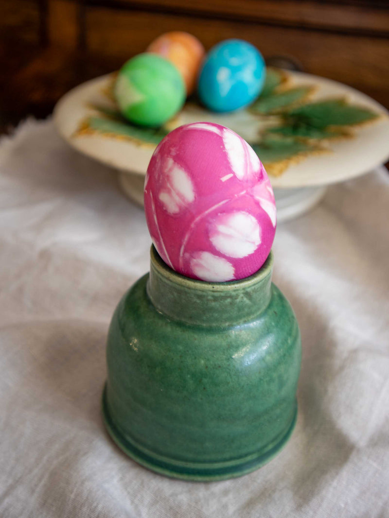 Decorative pink and white Easter egg in a green container on a white cloth with more eggs in the background.