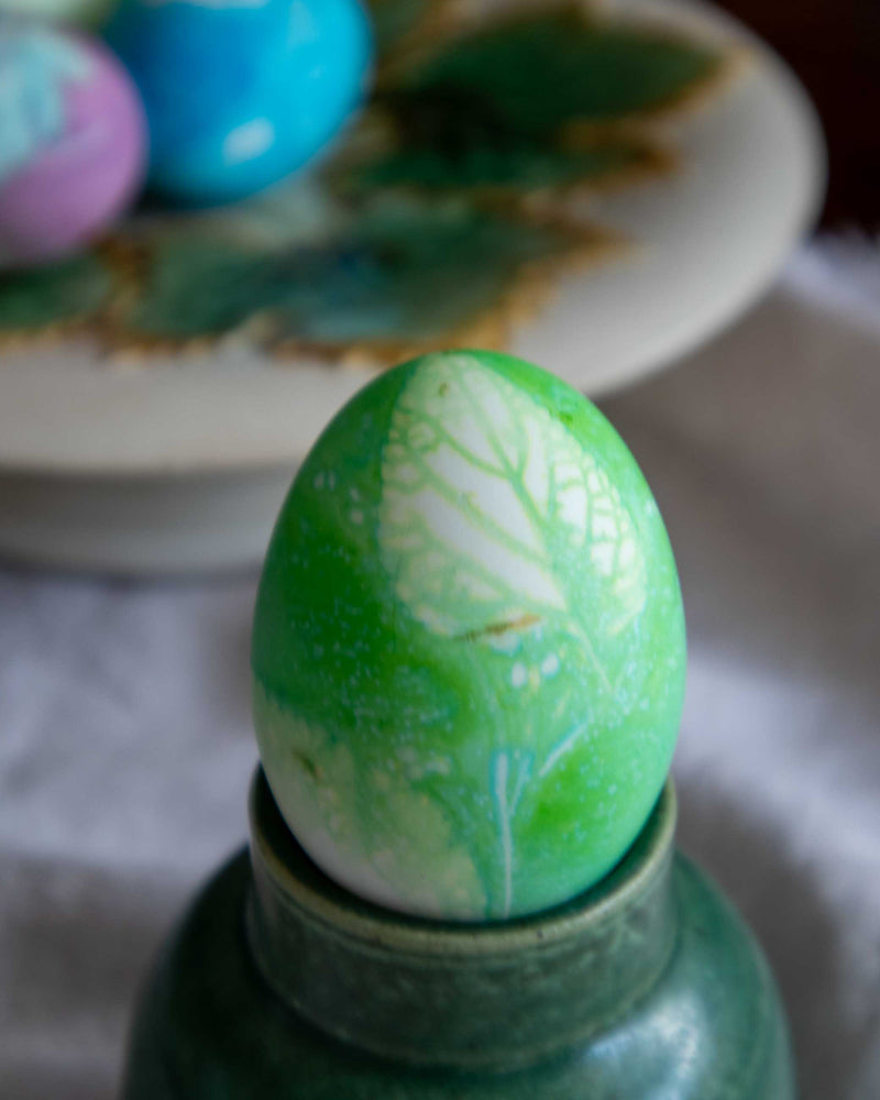 Green Easter egg with white leaf patterns on a stand, with blurred colorful eggs in the background.