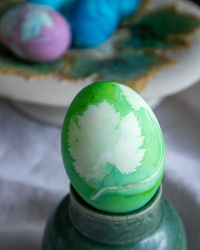 Green and white dyed Easter egg on a stand with blurred colorful eggs in the background.