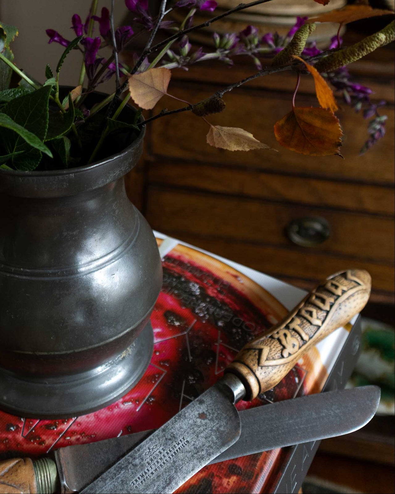 Decorative still life with a metal pitcher, purple flowers, and green leaves on a wooden surface.