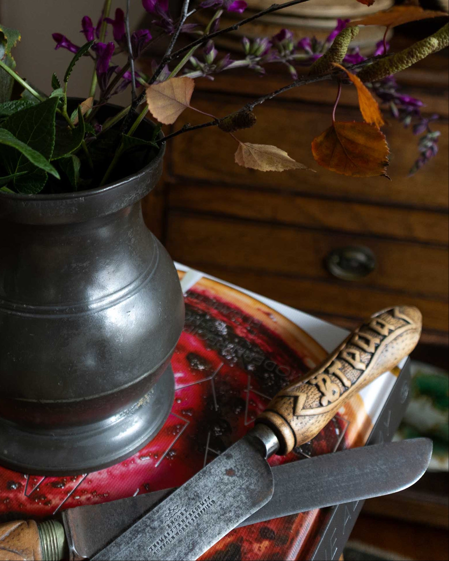 Decorative still life with a metal pitcher, purple flowers, and green leaves on a wooden surface.