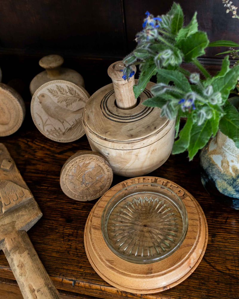 Wooden butter dish with glass insert on a wooden surface, surrounded by plants and butter prints and moulds.