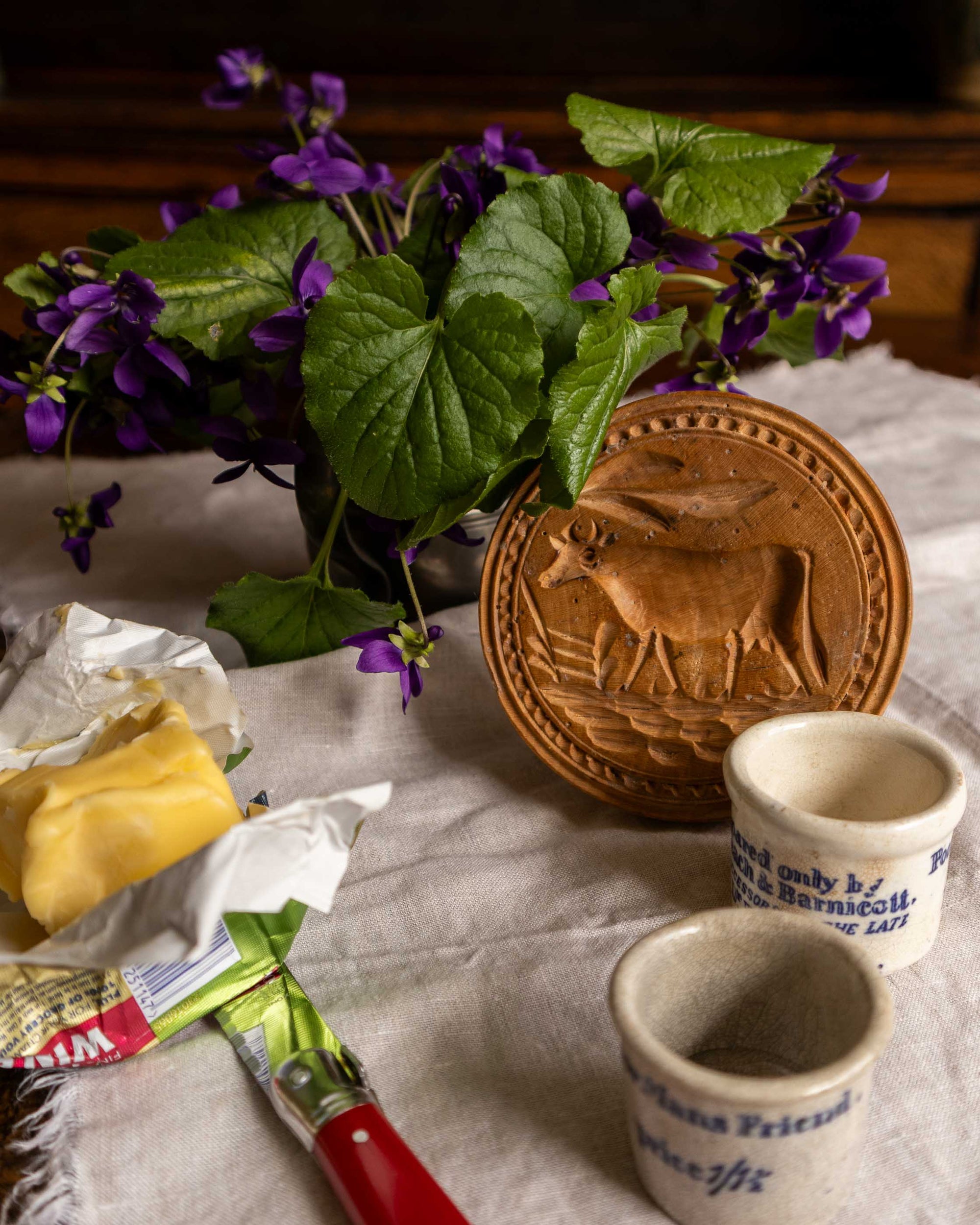 Wooden butter stamp with a cow design, purple flowers, and ceramic pots on a textured surface.
