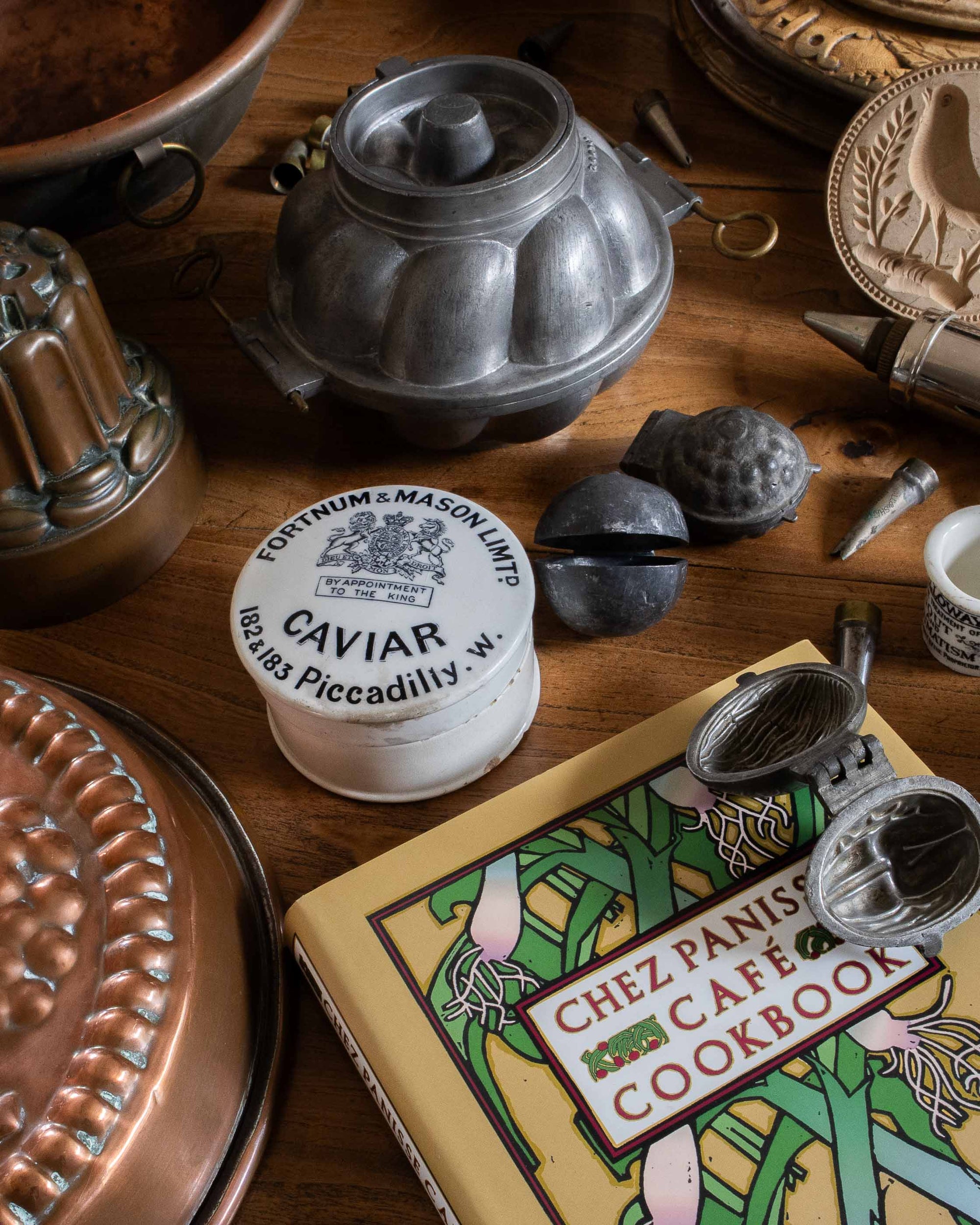 Vintage kitchen items including a caviar container, cookbooks, and copper and pewter moulds on a wooden surface.