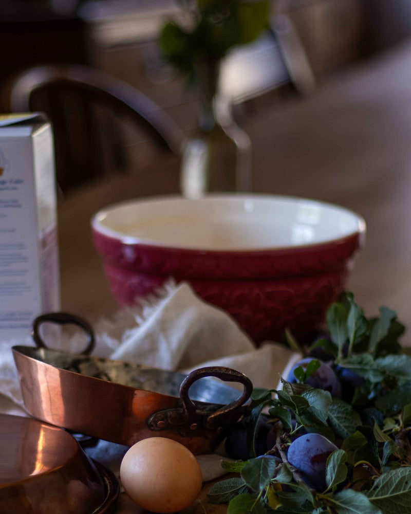 Red ceramic bowl on a table with a copper pan, egg, and plums.