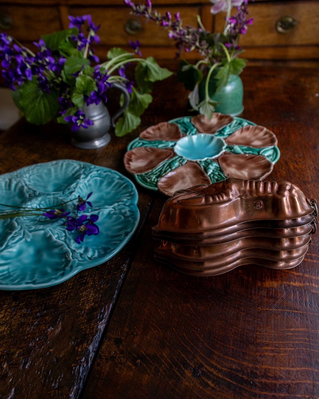 Ceramic majolica oyster plate and copper moulds and blue oyster plate on a wooden surface with purple flowers.