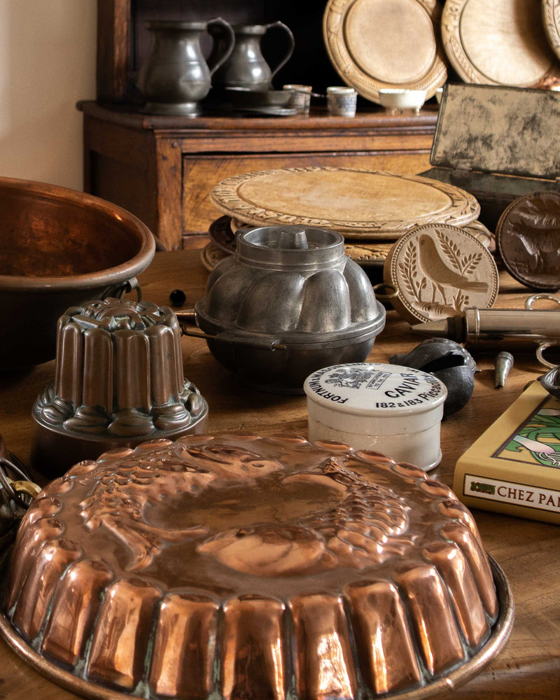 Collection of vintage kitchenware including copper moulds, metal pots, and wooden breadboards on a wooden surface.