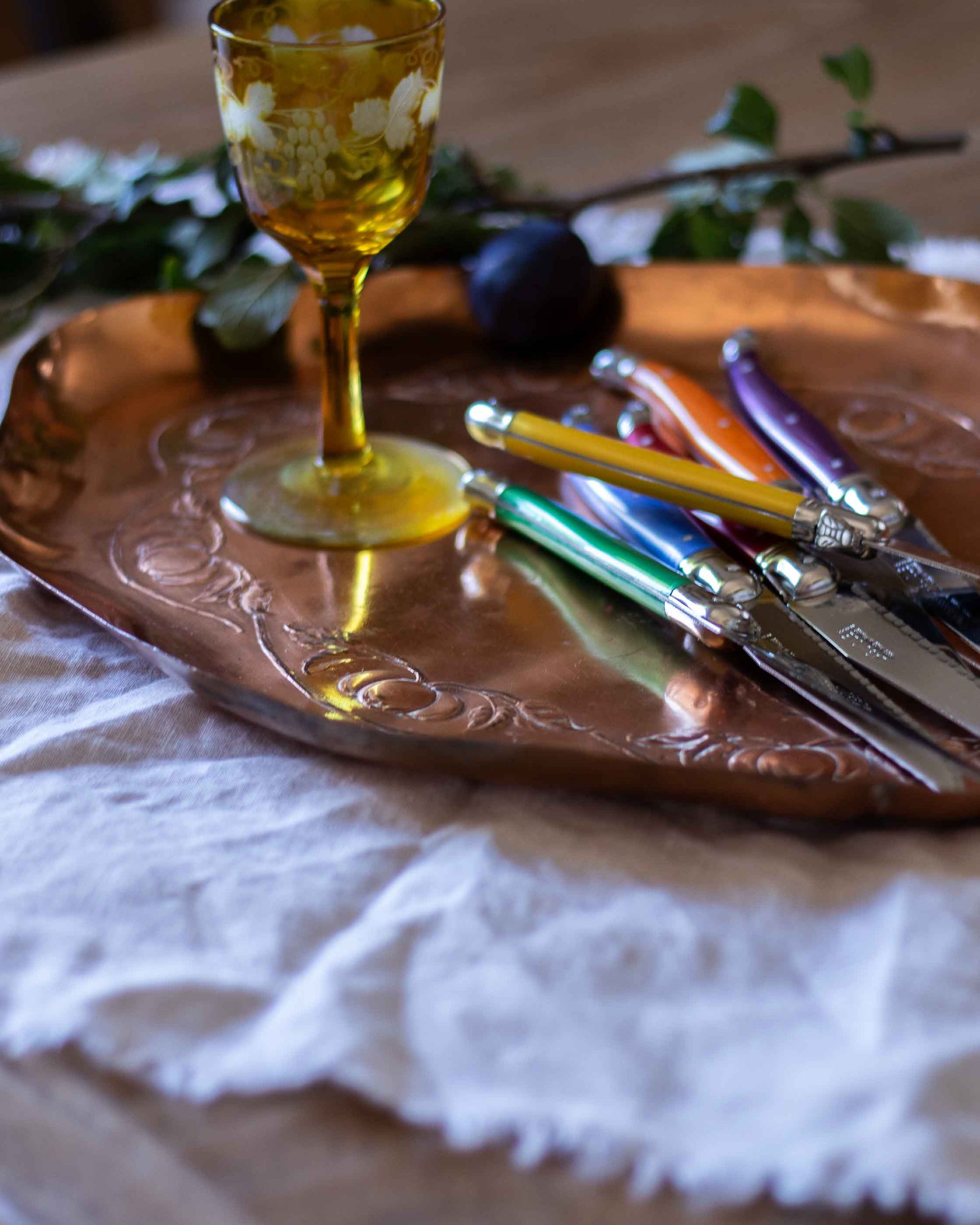 Decorative glass and colourful cutlery on a copper tray with a white cloth underneath.