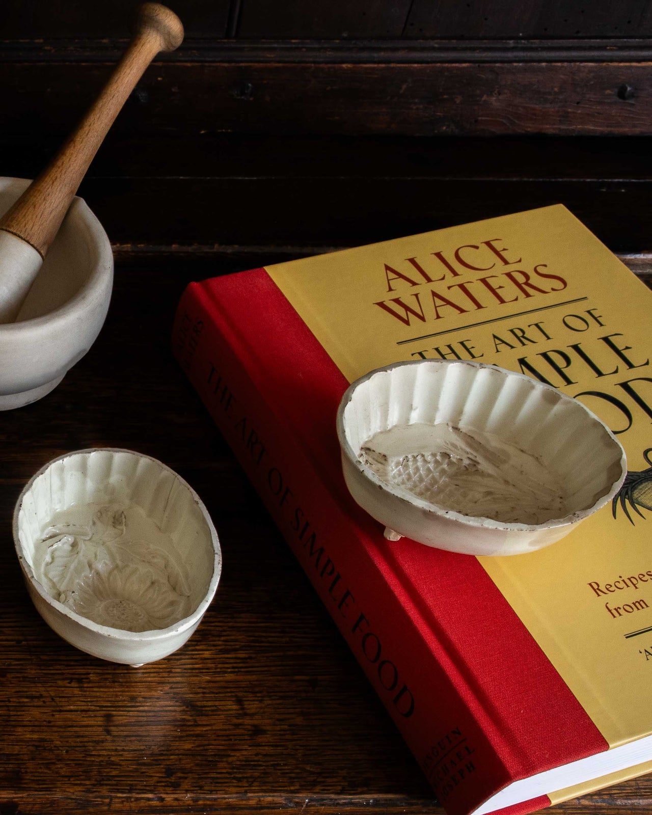Book titled 'The Art of Simple Cooking' by Alice Waters with small white moulds on a wooden surface.