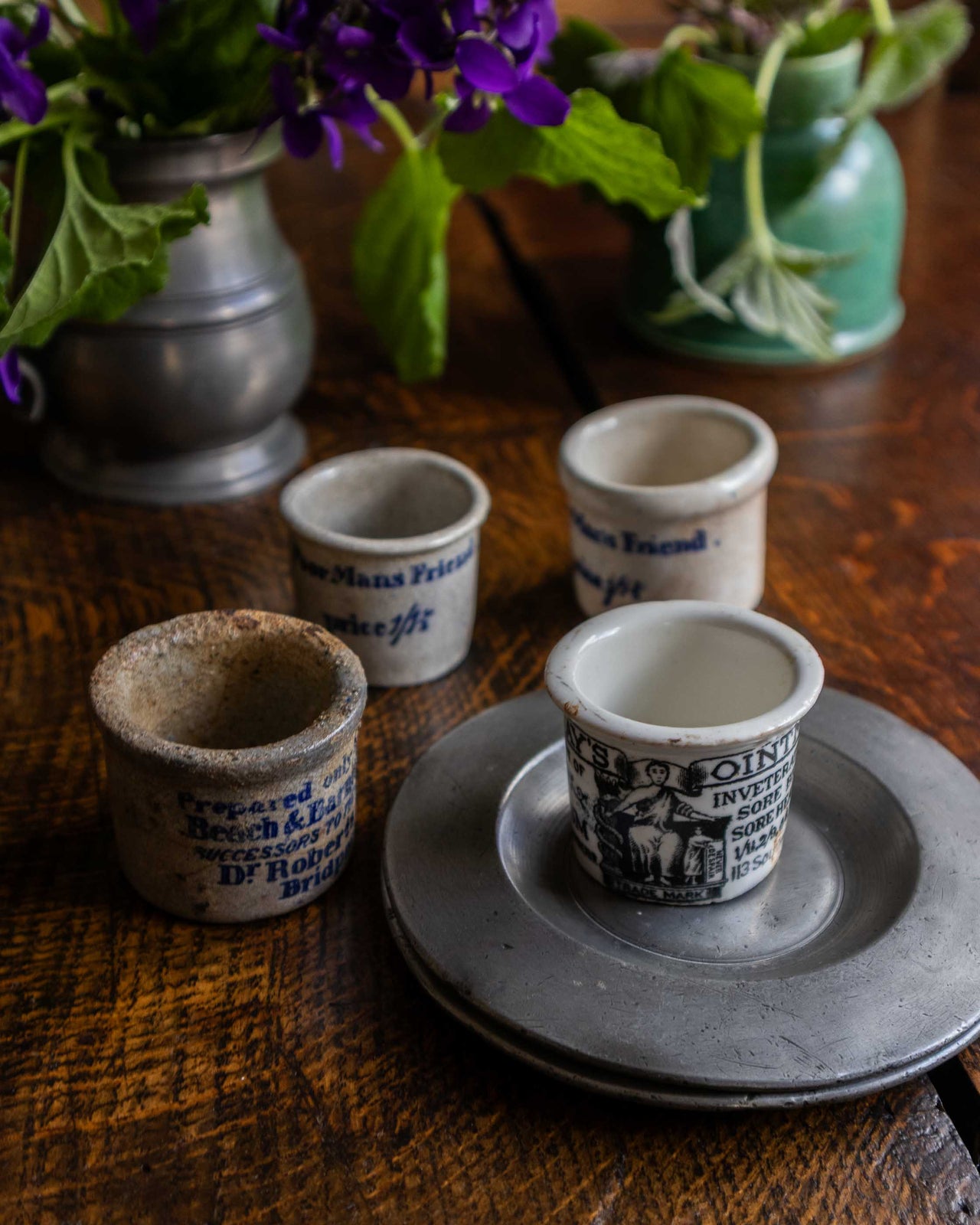 Set of small ointment pots with text on a wooden surface with plants in the background