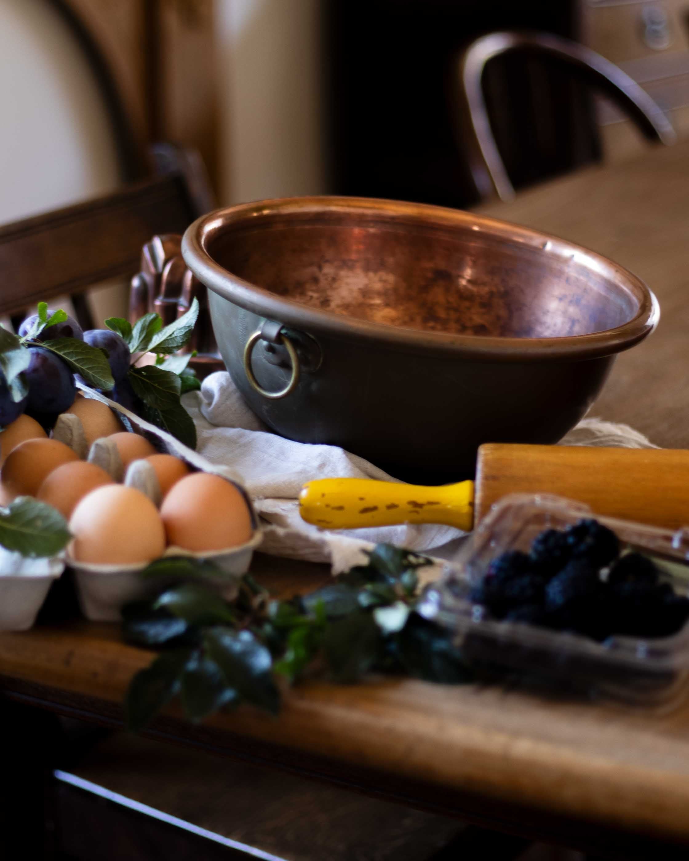 Copper bowl on a wooden table with eggs, plums, and a rolling pin.