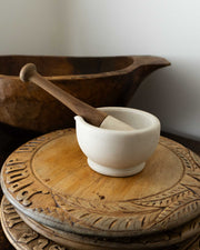 White mortar and pestle on a wooden breadboards with a wooden bowl in the background.