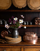 Vintage kitchenware including a copper jelly mould, pewter pitchers, and wooden breadboards on a wooden shelf.