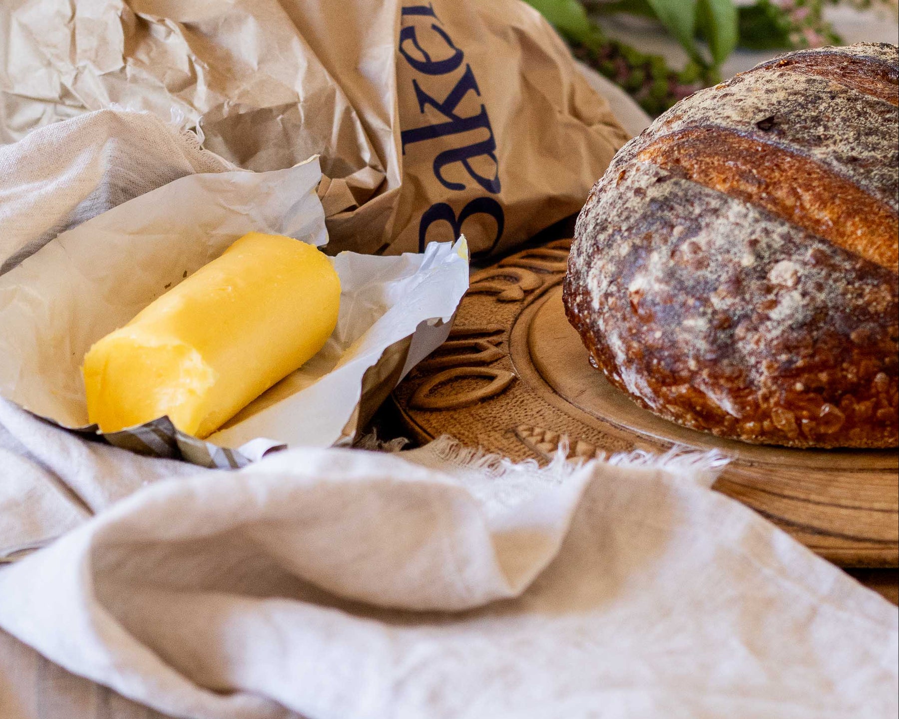 Butter stick on a wooden board with a loaf of bread and a Baker's paper bag in the background.