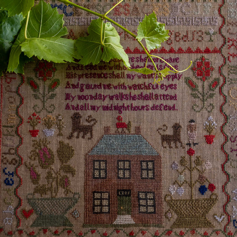 Embroidered antique sampler with house design and text, featuring leaves on top.