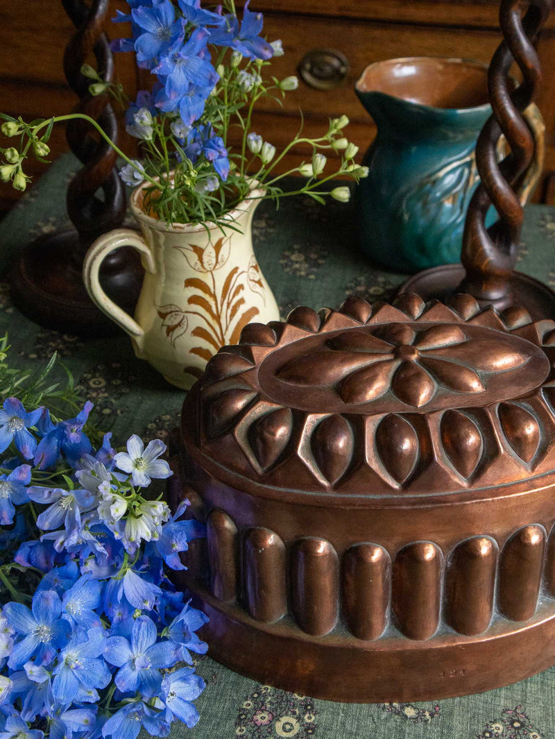 Decorative copper mould on a table with blue flowers and a pitcher.