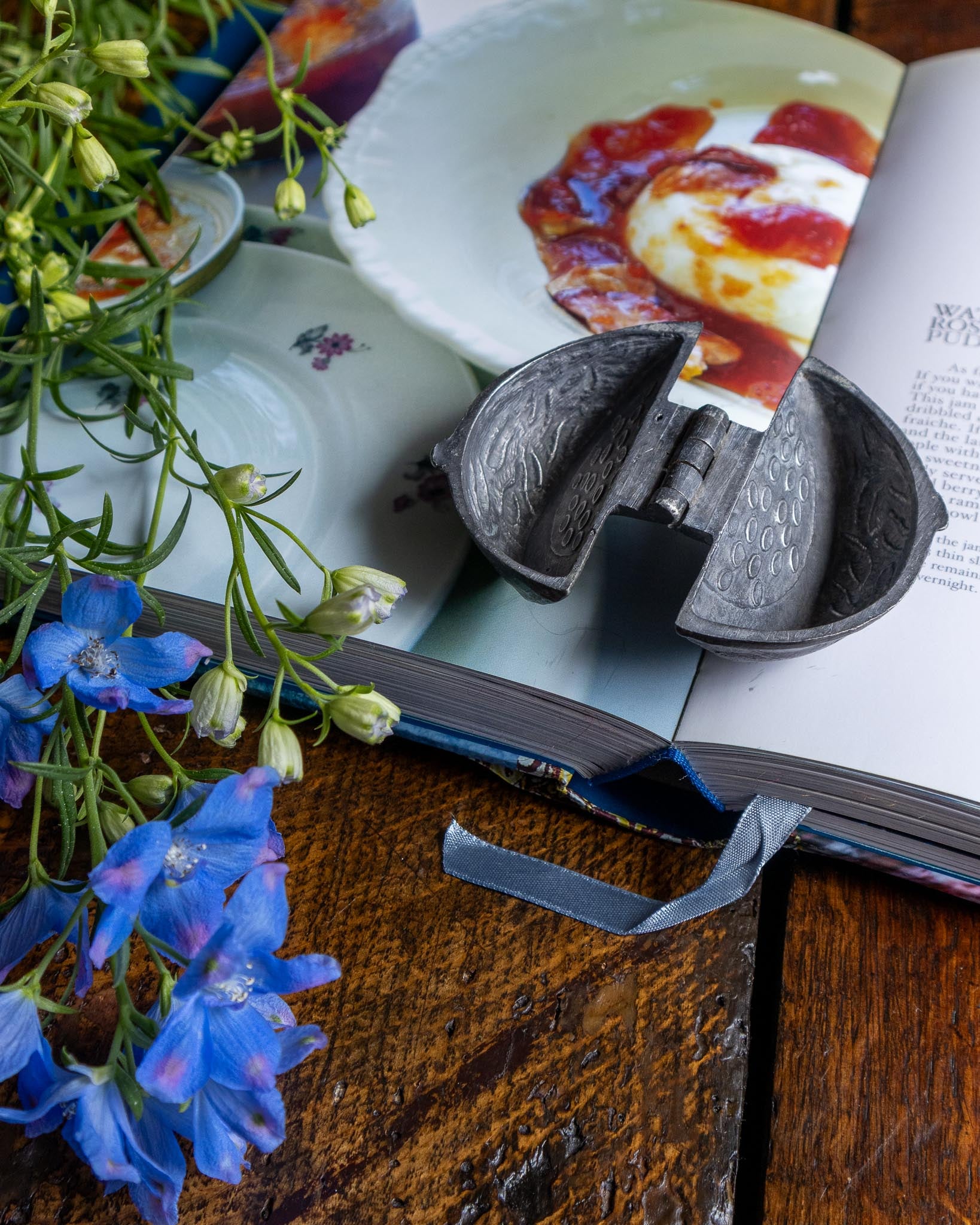 Decorative items including a pewter mould, blue flowers, and an open book on a wooden surface.