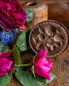 Wooden butter stamp with floral design surrounded by pink roses on a wooden surface