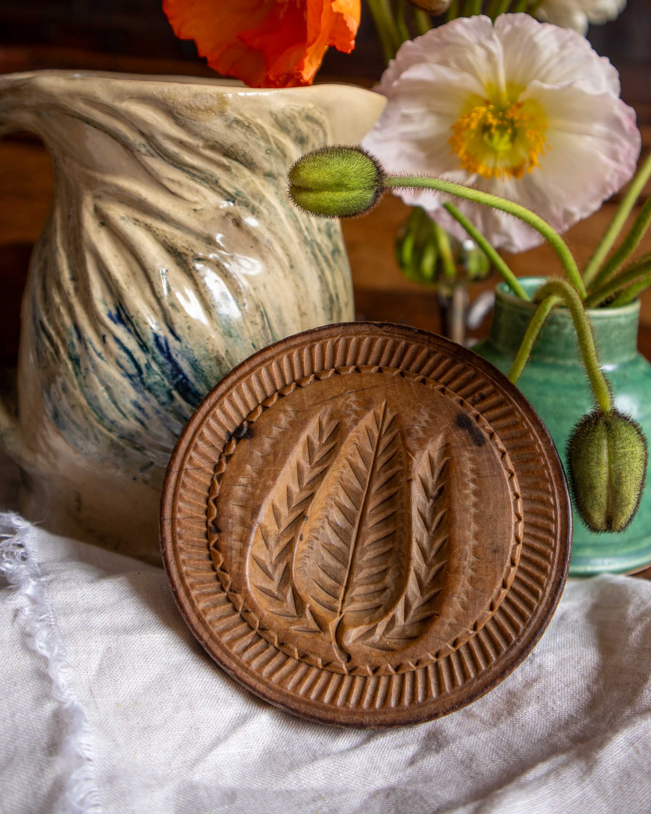 Wooden butter print with leaf design on a white cloth with a ceramic pitcher and flowers in the background