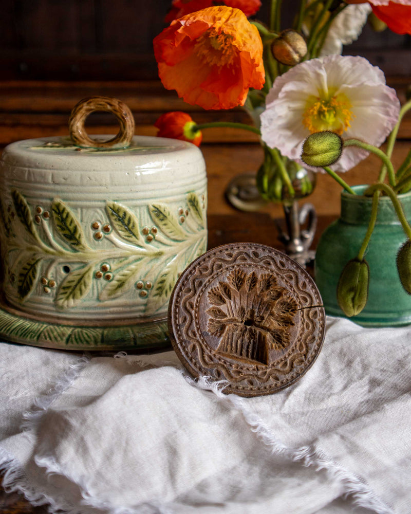 Decorative ceramic cheese dish with floral patterns, wooden butter stamp, and flowers in the background.