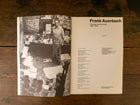 Open book showing a black and white photo of Frank Auerbach in a studio and text on a wooden surface