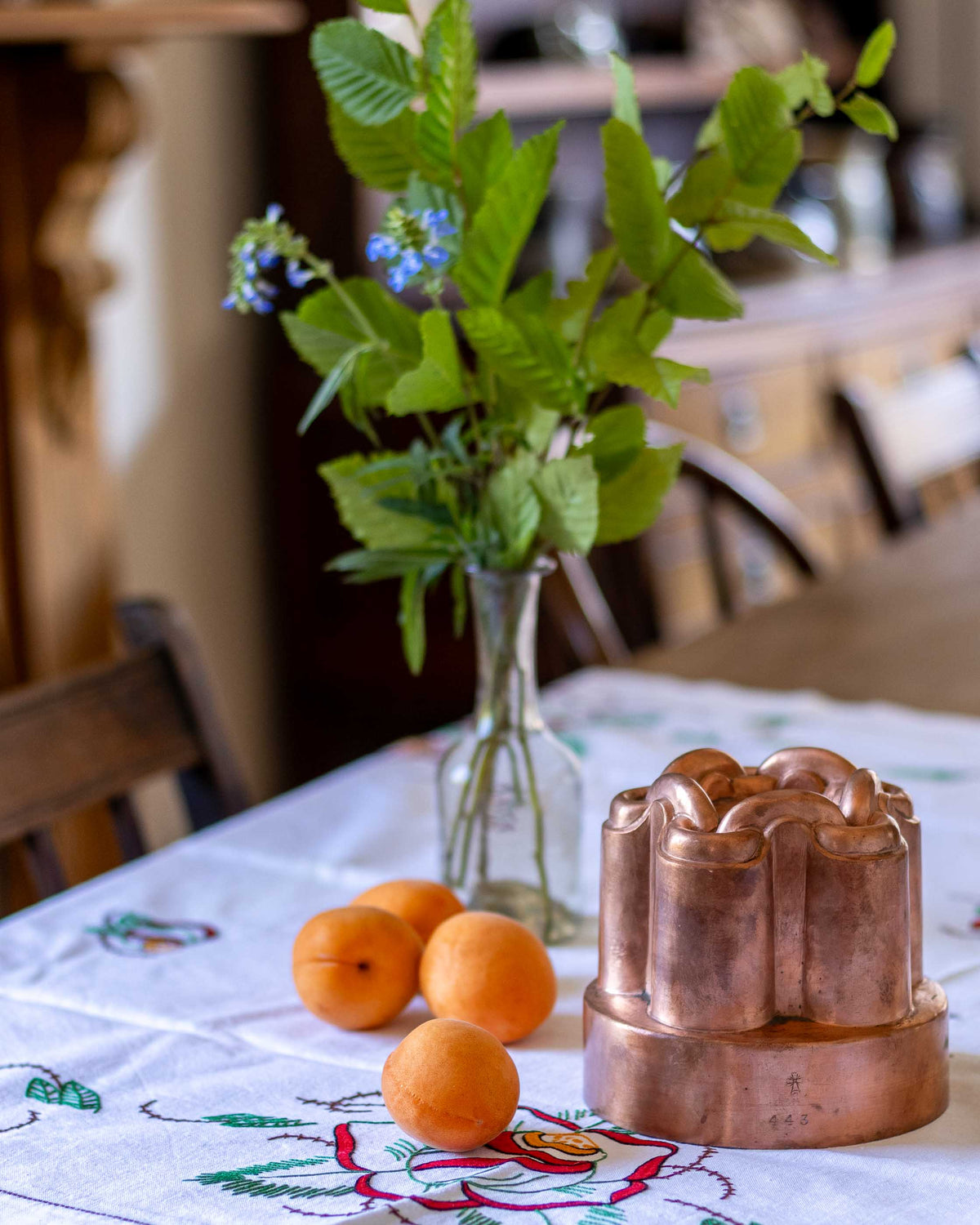 Copper mold with apricots and a vase of flowers on a tablecloth.